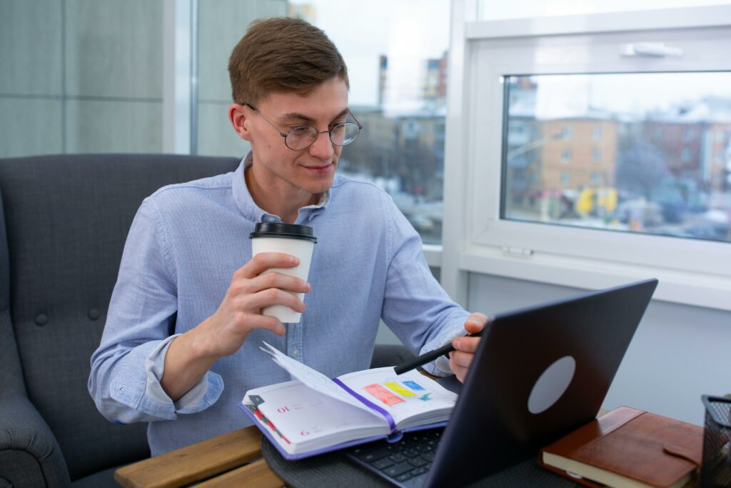 Business professional sipping coffee while working on a laptop with a planner at an office.